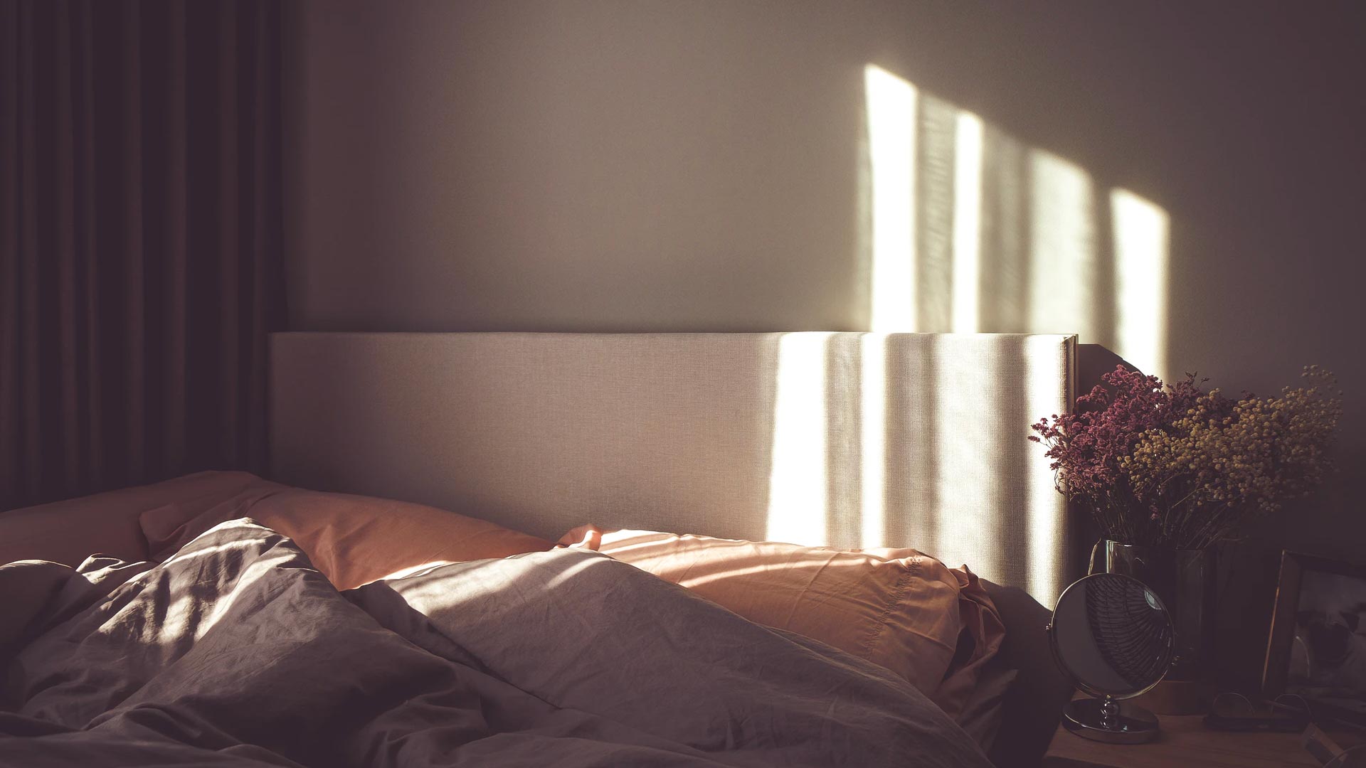 A dark lit room and unmade bed with some light coming in between the curtains