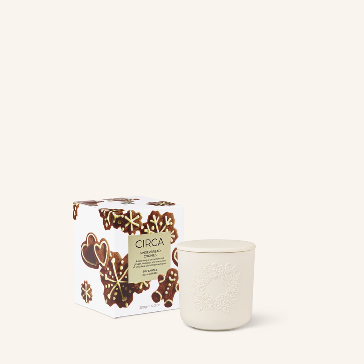 White ceramic candle with an embossed wreath and a lid. The box is on an angle. The candle packaging features gingerbread cookies and gingerbread men on a white background.
