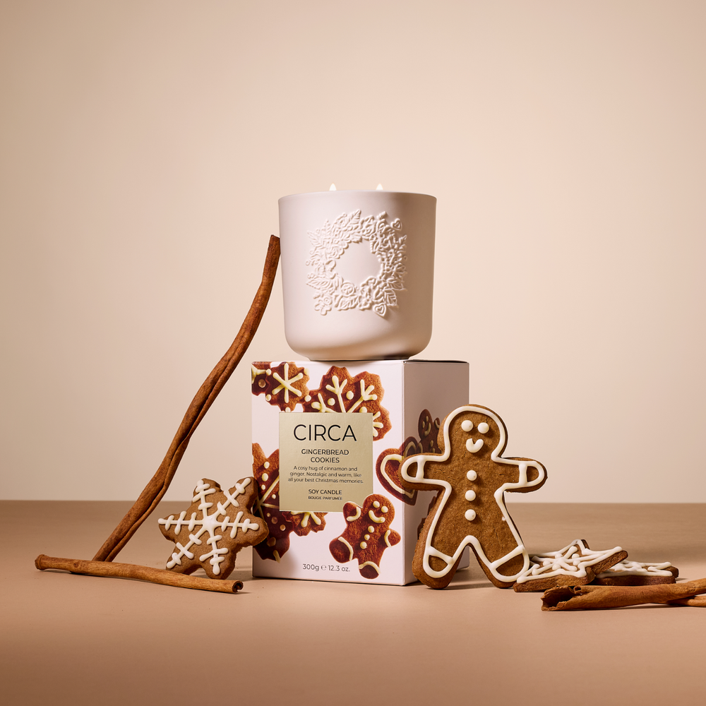 Circa ceramic white candle with gingerbread-themed packaging, cookies and cinnamon sticks on a beige background.