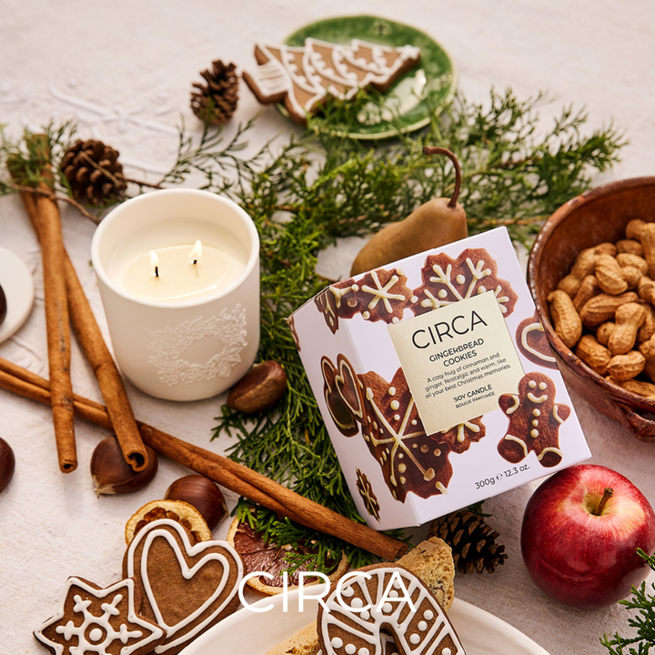 Circa white ceramic candle burning. Surrounded by gingerbread cookies packaging, cinnamon sticks, greenery and apples on a decorative surface.