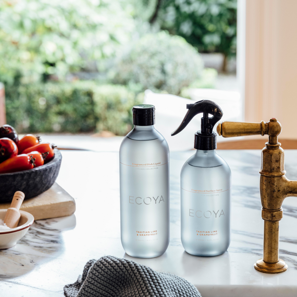 Two ECOYA bottles, Dishwashing Liquid and Surface Spray in Tahitian Lime & Grapefruit fragrance, on a kitchen counter with a faucet and vegetables in the background.
