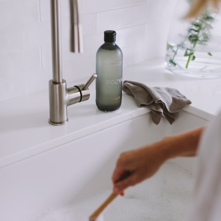 Person cleaning dishes in a kitchen sink with a bottle of ECOYA Tahitian Lime & Grapefruit dishwashing liquid and a cloth.